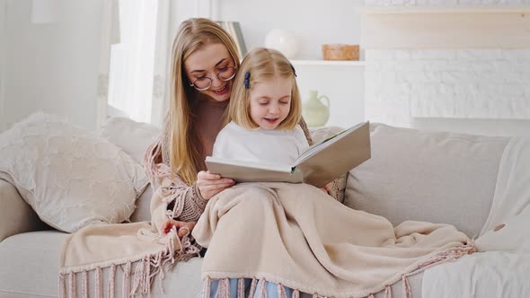 Young Mom Blonde Mother Sits with Little Daughter Baby Girl on Couch Covered By Warm Blanket Reading alt
