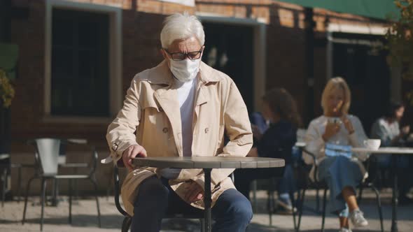 Elegan Aged Man Sitting Down at Table in Street Cafe Putting Off Safety Mask alt