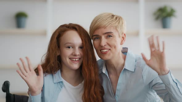 Little Handicapped Redhead Girl Sitting in Wheelchair Embracing with Her Mother and Waving Hand to alt