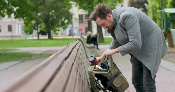 Young Man with Mustaches and a Beard is Searching for Something in His Backpack Placed on the Wooden alt
