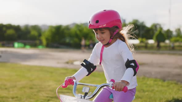 Cute Light Hair Little Girl in Pink Helmet in Elbow and Knee Pads Rides a Bicycle Smiles and Feels
