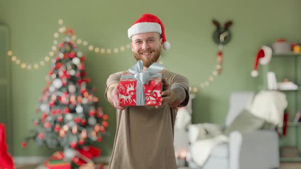 Young Handsome Bearded Tattooed Guy in Green T-shirt with Red Christmas Hat Is Giving a Gift To the alt