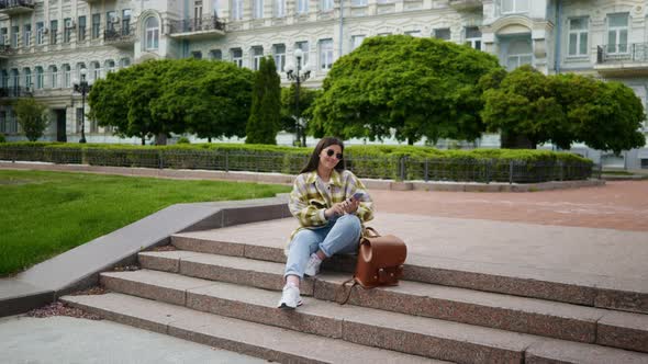 A Girl Sits in the Park on the Stairs Listening to Music and Texting on Her Phone