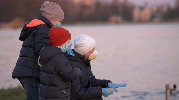 Mother, son and daughter in medical masks feed ducks in the Park on the lake.