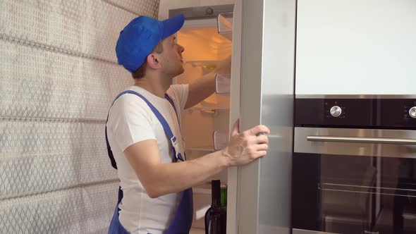 a young repairman in protective overalls moves the refrigerator in the kitchen. alt