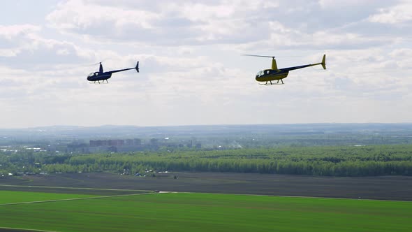 Two Bright Colored Robinson Helicopters Fly Over Green Fields Against Sky with Clouds on Sunny Day