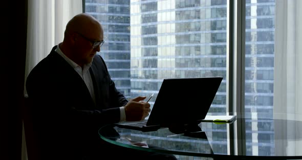 Silhouette of a Businessman in Front of the Window Sits at a Table with a Laptop and Goes on a Video alt