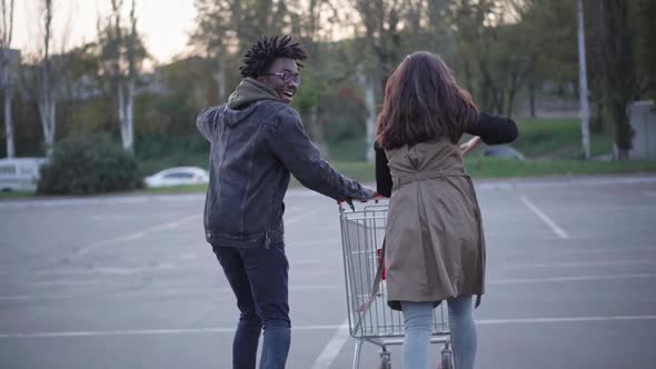 Back View of Cheerful Interracial Young Couple Running with Shopping Cart on Parking Lot alt