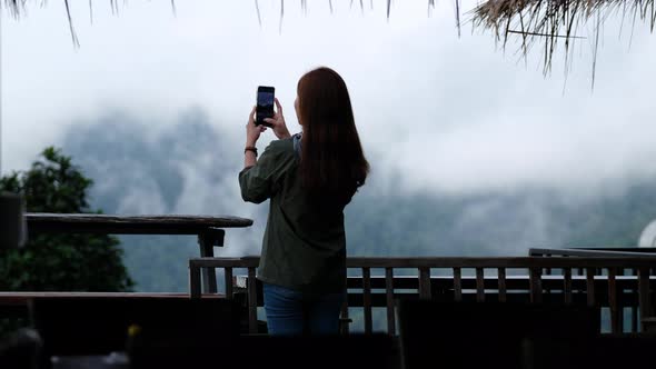 A female traveler using mobile phone to take photo of a beautiful foggy mountain and nature view