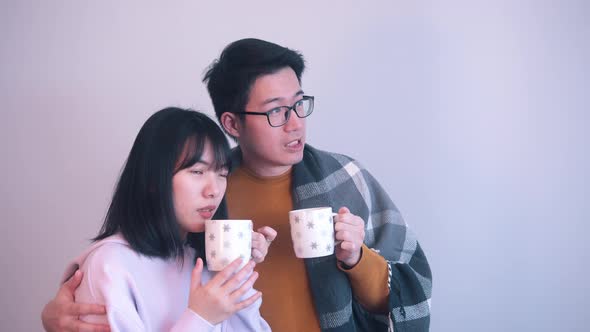 Young Asian Couple Hugging Under the Blanket and Drinking Hot Beverage Indoors on Stormy Day alt