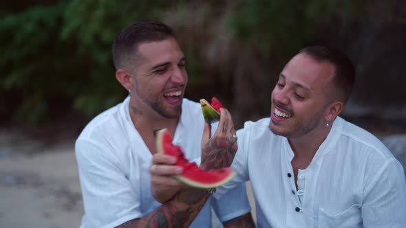 Diverse Gay Male Couple Eating Watermelon Together and Talking Sitting on the Rocky Beach alt
