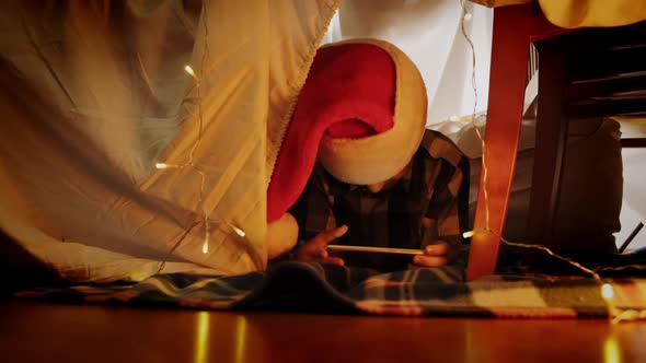 Kid in Santa Hat in DIY Kid's Tent Holding Smartphone Having Joyful Conversation alt