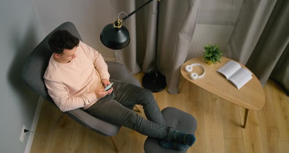 Top View of Man Using Smartphone App at Home at Night Sitting in Armchair