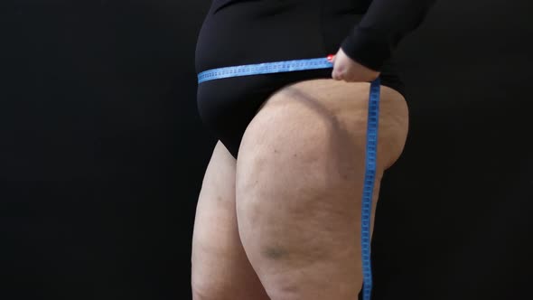 a Fat Woman with Cellulite Measures Herself with a Measuring Tape on a Black Background