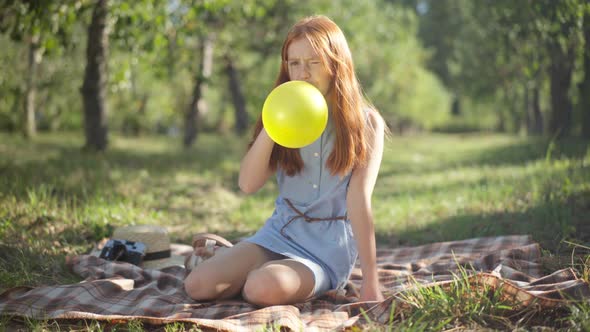 Wide Shot Portrait of Redhead Beautiful Teenage Girl Blowing Balloon Sitting on Blanket in Spring alt