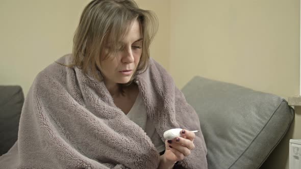 Young Woman Sits Wrapped in a Blanket and Measures the Temperature alt