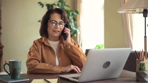 Woman Talking on Phone and Using Laptop at Table in Apartment Room alt