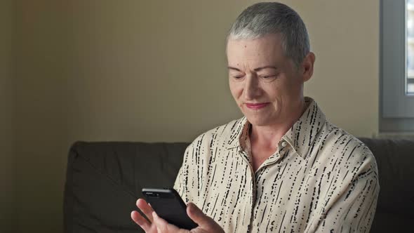 An Elderly Woman Looks at the Smartphone Screen and Smiles Sincerely