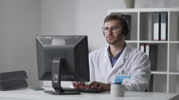 Male General Practitioner in White Coat Sitting at Desk in Doctor's Office and Scrolling Computer alt