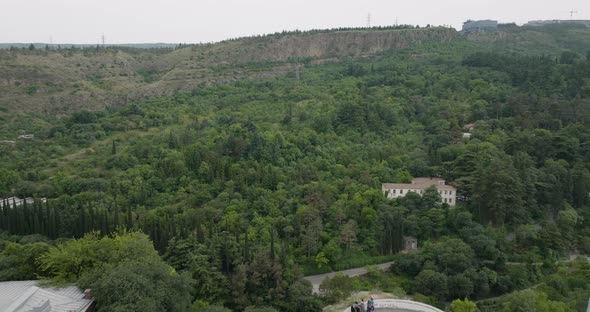 Dolly out aerial shot of the Tbilisi forest, revealing the Kartlis Deda statue. alt