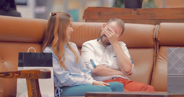 Man and a Girl are Sitting on a Sofa in a Mall Talking and Laughing alt