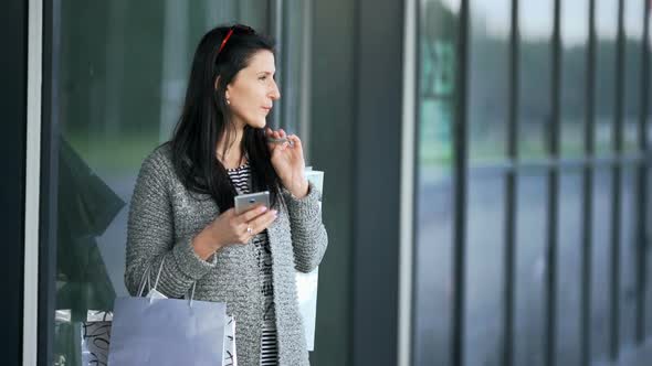 Beautiful Young Fashion Woman with Shopping Bags Near Shop Window Calling Smart Phone alt