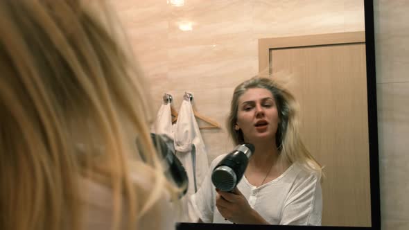 Young Woman Dances and Sings in a Working Hair Dryer in Front of a Mirror Standing in the Bathroom