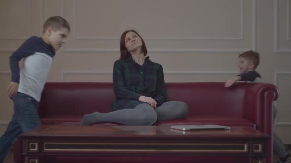 Exhausted young mother sitting on home red couch while two loud kids running around  alt