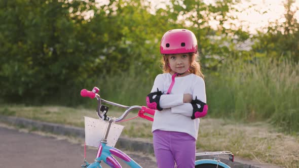 Cute Light Hair Little Girl in Pink Helmet in Elbow and Knee Pads Folds Her Hands Next to Bicycle in