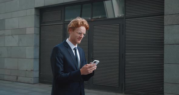 Young Caucasian Male Businessman Typing on Smartphone While Walking Along Modern Business Building alt