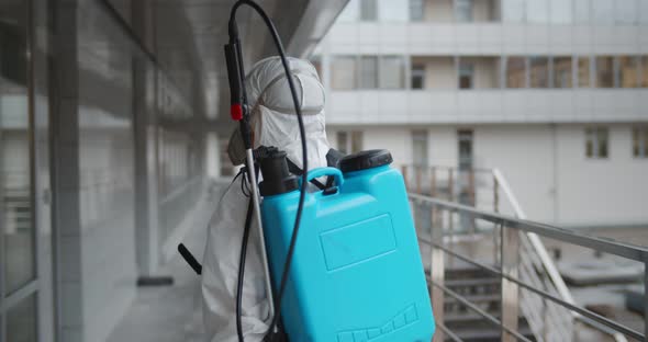 Back View of Health Worker in Sterile Clothing Spraying Chemical Outside Office Building alt