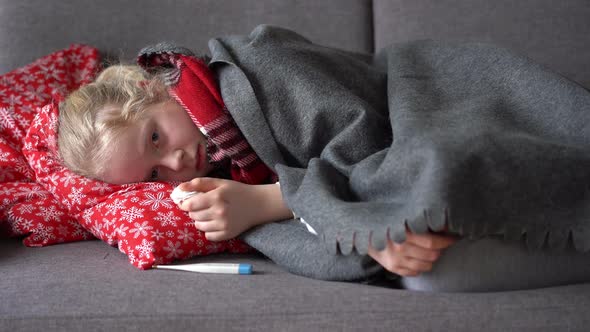 Cold Caucasian Girl at Home. Portrait of a Sick Child in a Scarf and Plaid on the Sofa