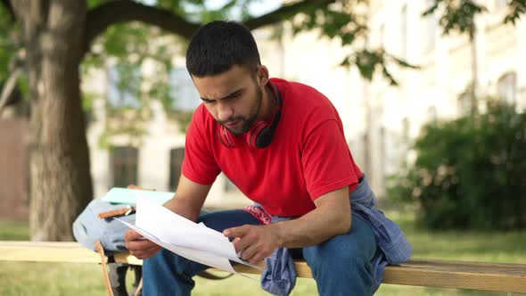 Exhausted Young Male Student with Paperwork Holding Head in Hands Sitting on Bench Outdoors on Sunny alt