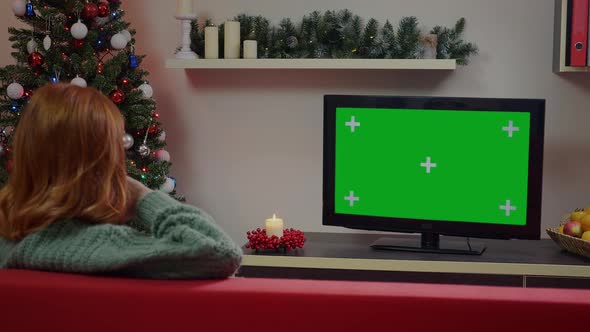 A red-haired girl watching Green Screen Mockup TV in a Chrismas decorated living room. alt