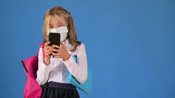 Portrait of a schoolgirl with a briefcase in a protective mask on her face uses a smartphone alt