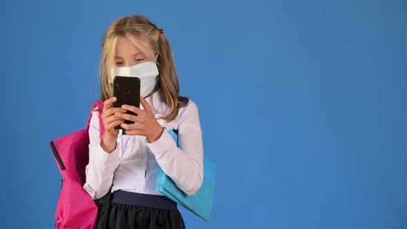 Portrait of a schoolgirl with a briefcase in a protective mask on her face uses a smartphone