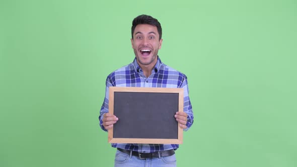 Happy Young Hispanic Man Holding Blackboard and Looking Surprised alt