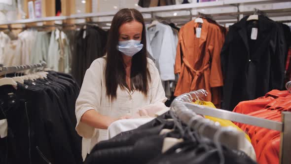 A Woman in a Medical Mask in a Clothing Store Chooses New Fashionable Things for Herself During the