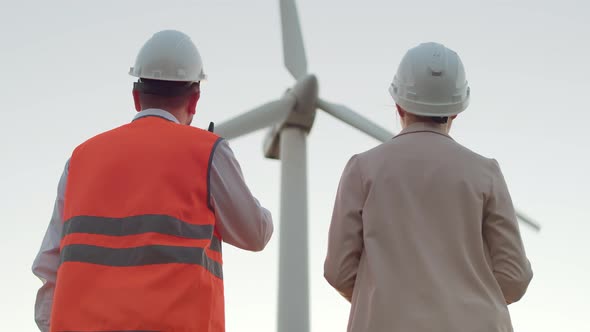 View From the Back of a Man in a Special Suit and a Girl in a Hard Hat Looking at a Windmill, a Man alt