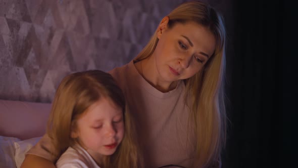 Woman Looks at Daughter Reading Book on Bed at Night alt