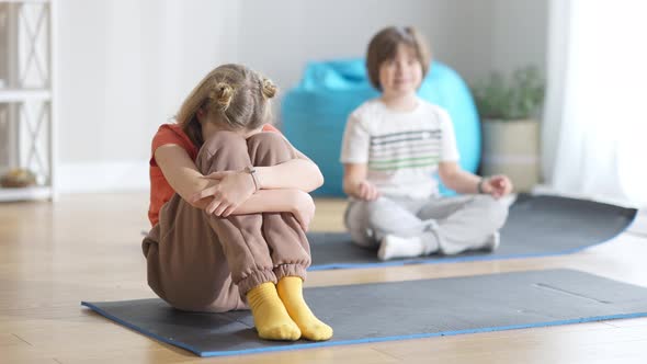 Sad Little Girl Hugging Knees Sitting on Exercise Mat As Blurred Confident Boy in Lotus Pose alt