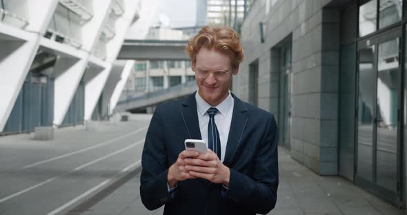 Redheaded Smiling Businessman in Eyeglasses and Formal Suit Stand in the City Center Street Uses alt