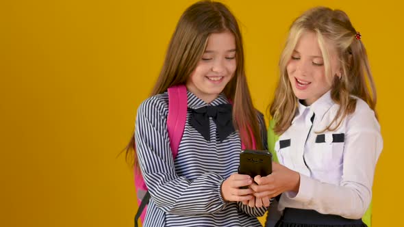 Two schoolgirls friends in school uniforms with backpacks use a smartphone
