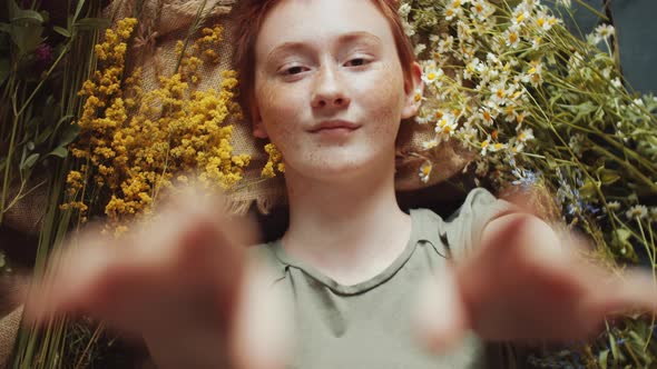 Redhead Girl Reaching Out Hands to Camera while Posing among Flowers alt