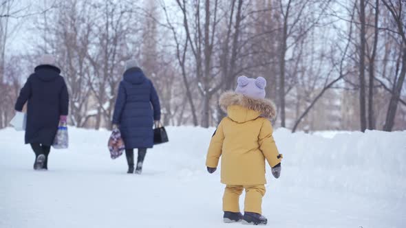 toddler 12-17 months old in yellow winter clothes looks after two middle-aged women alt