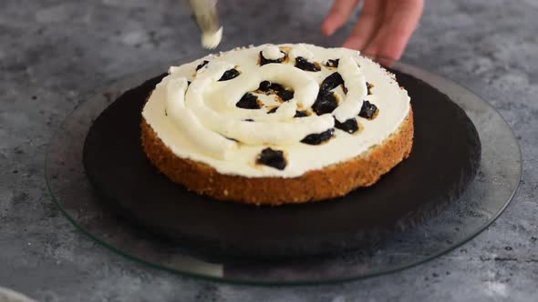 Caucasian woman's hand decorates cake squeezes cream out of pastry bag on rotating round stand. alt