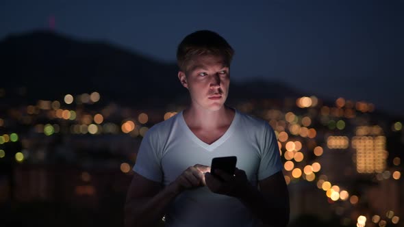 Young Happy Tourist Man Using Phone Against View Of The City Lights At Night alt