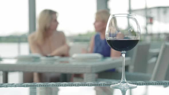 Glass with Red Wine Standing at the Table in Outdoor Cafe and Two Blurred Blond Caucasian Women alt