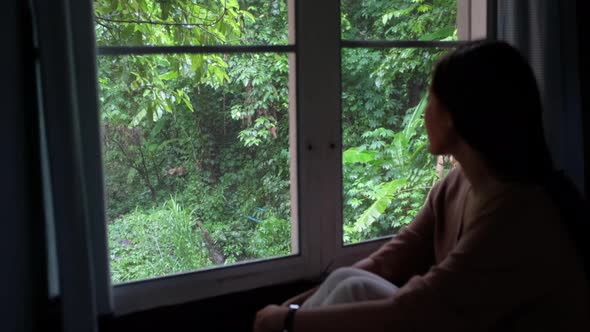 A woman sitting in the house and looking at a beautiful nature outside the window while it's raining