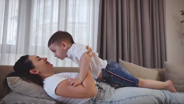 Happy Healthy Family Young Mom Raising Cute Little Child Son Up Playing on Sofa at Home, Mom Kisses alt